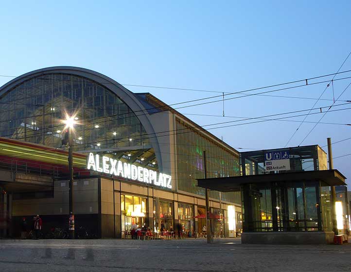 Nahaufnahme der bogenförmigen, beleuchteten Glasfassade des Bahnhofs Alexanderplatz bei Dämmerung, mit leuchtendem Schriftzug am Eingang.